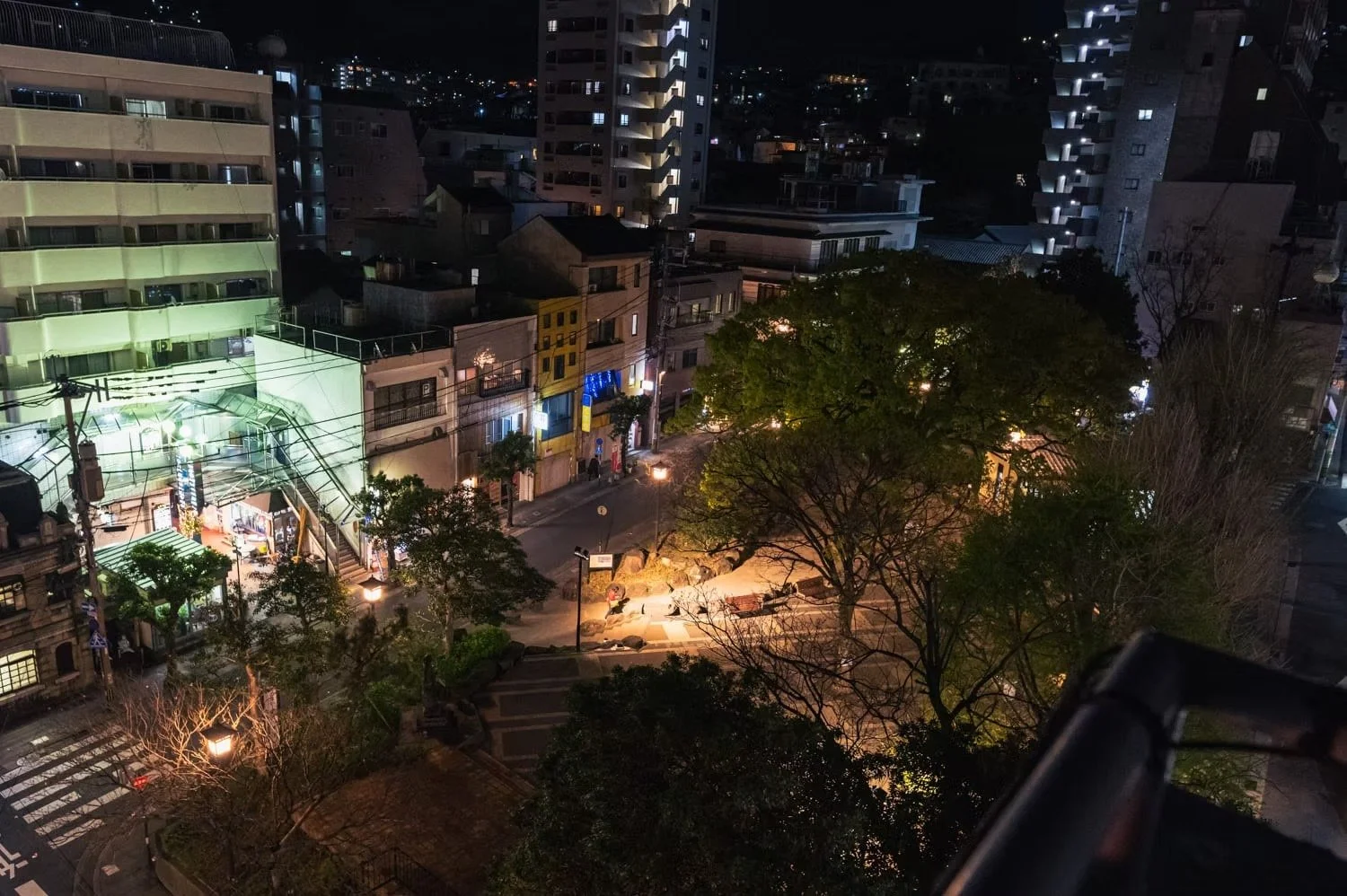 Nighttime city street view with buildings, trees, street lights, and illuminated storefronts.