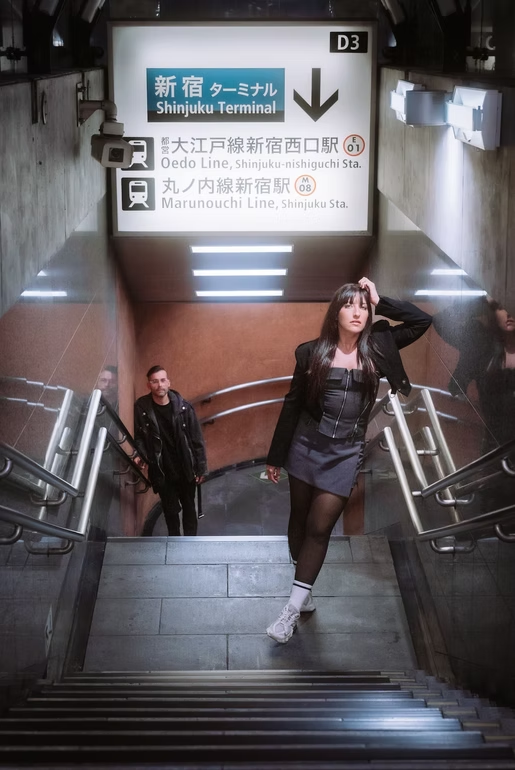 A woman with long dark hair wearing a black jacket, black dress, and white sneakers with black tights, standing on stairs in Shinjuku Terminal in Tokyo, Japan, with two men in the background. Sign above indicates directions to the Oedo Line and Marunouchi Line subway stations.