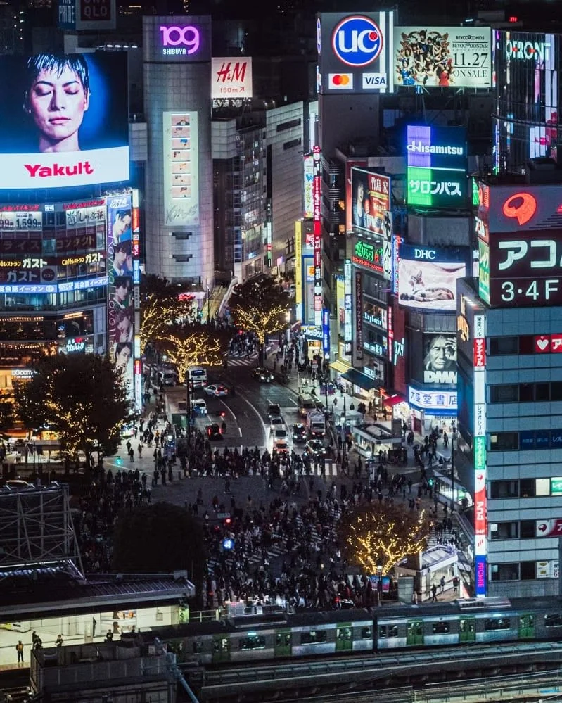 A bustling city intersection at night with crowded crosswalks, tall buildings illuminated by neon signs and billboards advertising various brands and products, including electronics, fashion, and entertainment, with a train passing at the bottom of t