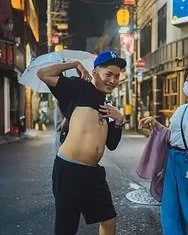 A young man in a blue cap lifts his shirt and poses playfully on a Tokyo street at night, holding an umbrella, while a woman walks past him.