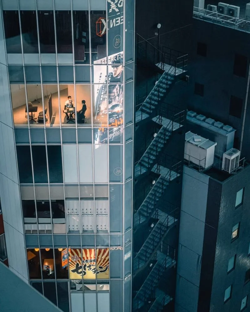 Office building with glass windows, showing two people in a meeting, a large outdoor poster, and exterior fire escape stairs.