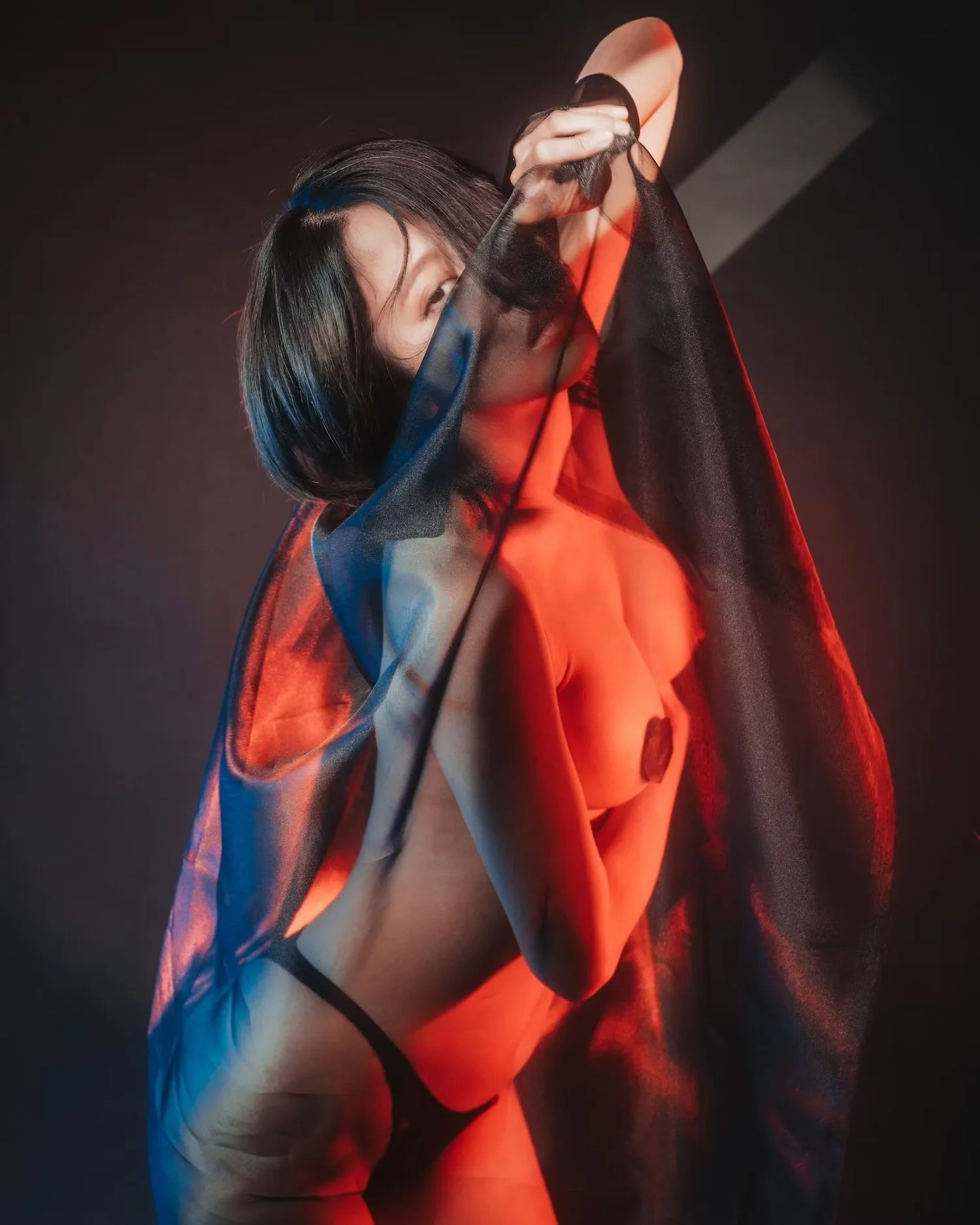 A woman with dark hair partially covered by a sheer black fabric, wearing a red top, and posing against a dark background with red and blue lighting.