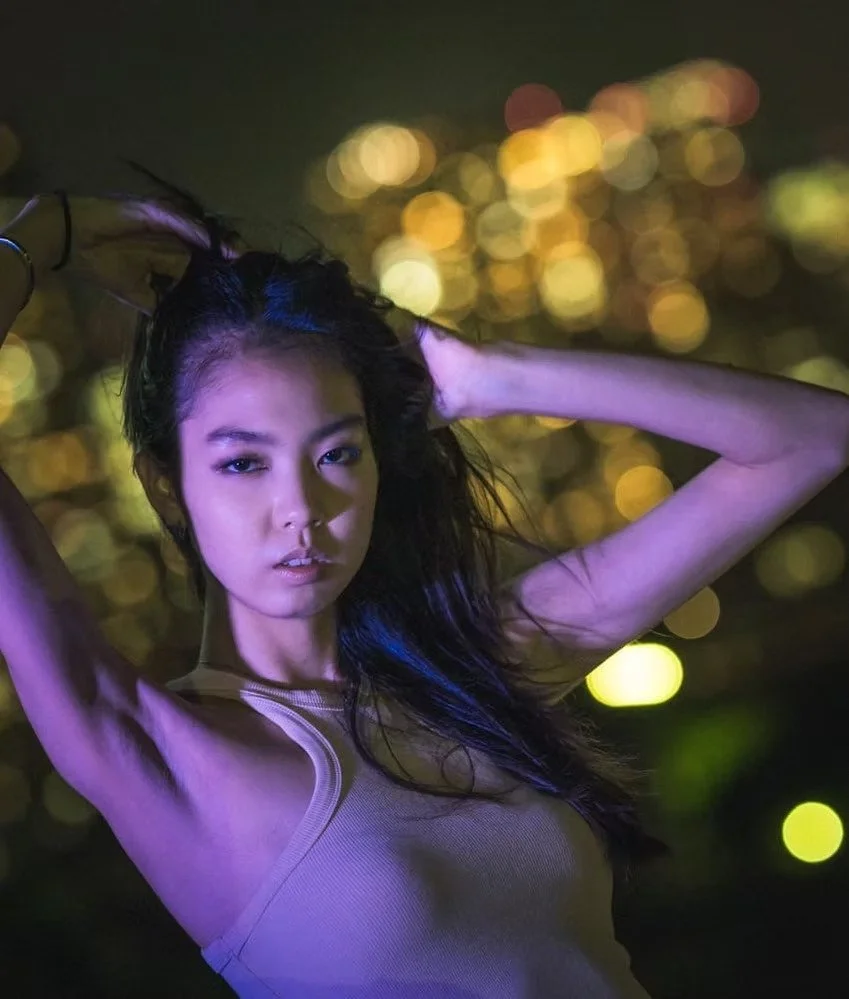A woman with long dark hair posing against a blurred city lights background at night, holding her hair with one hand and looking at the camera.