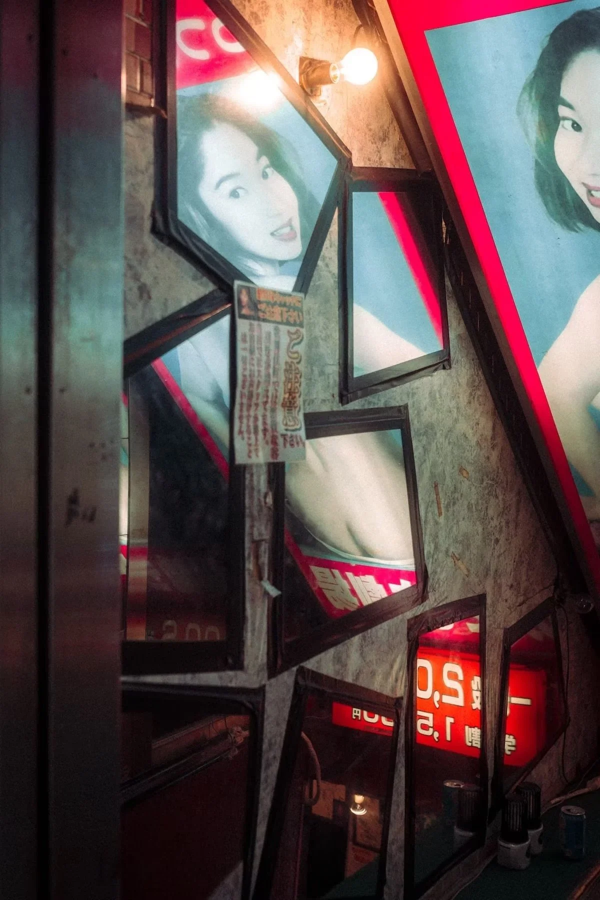 Photographs of women on illuminated posters or signs, displayed on a wall in a dimly lit area, with some frames leaning against the wall. Red light district in Tokyo.