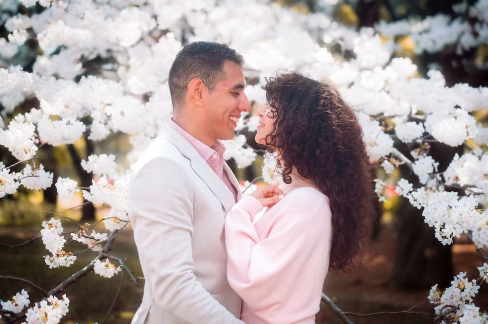 A couple stands close together in a park, gazing at each other with cherry blossoms blooming in the background. The scene captures a romantic and serene moment.