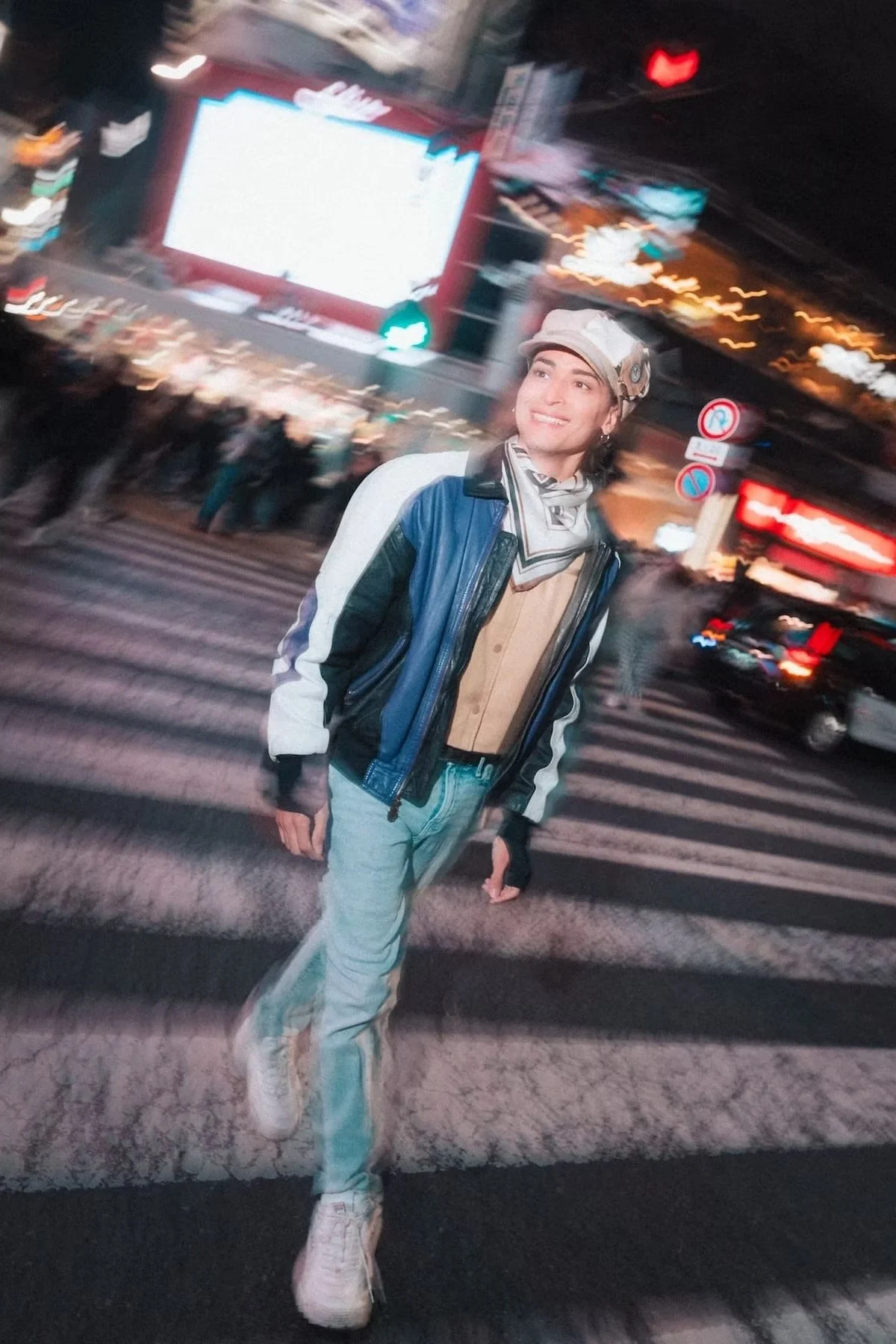 A young person confidently walks across a busy crosswalk at night in a vibrant city, with neon lights illuminating the scene. The motion blur captures the energy and fast pace of the urban environment, emphasizing the individual’s bold fashion style.