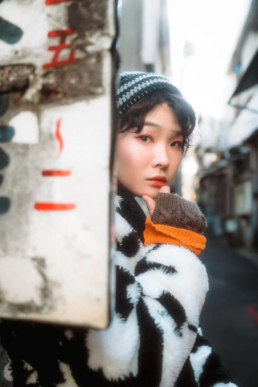 A young woman with short dark hair wearing a striped beanie and a black and white fuzzy sweater with orange cuffs, posing behind a wall in an outdoor alley with buildings and a reflective surface in the background.