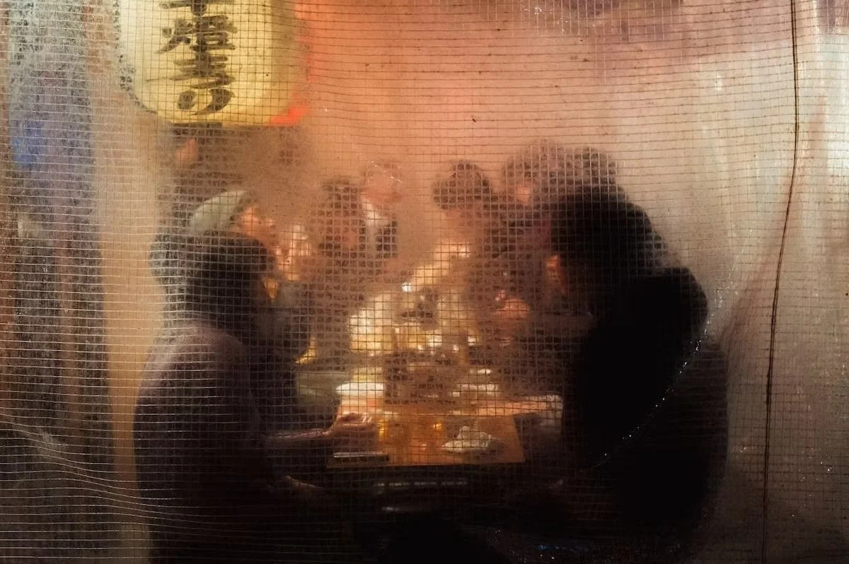 People cooking or eating at a table behind a mesh screen, likely at a restaurant. Izakaya night scenes shot by Tokyo street photographer Deniz Demir.
