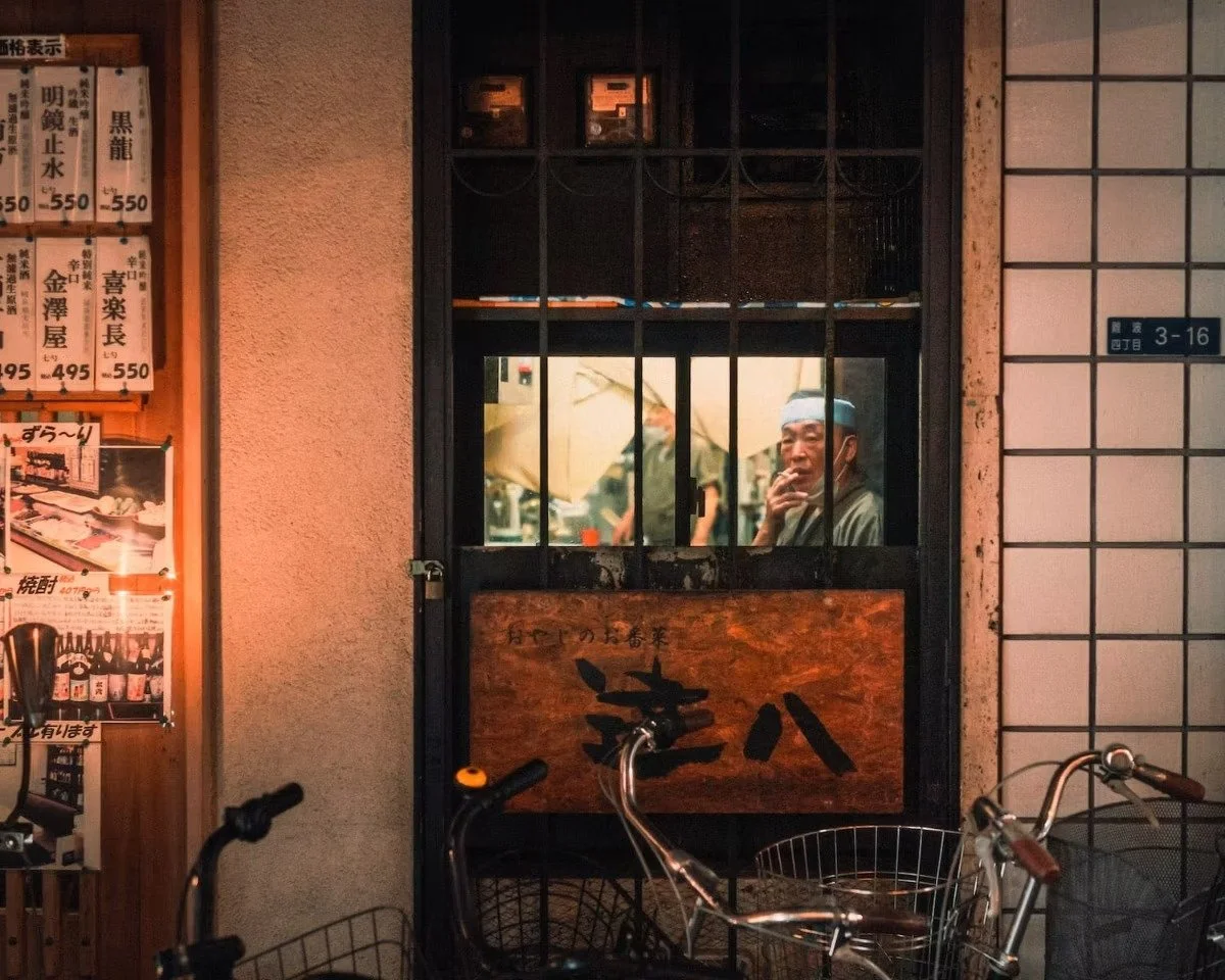 A street scene showing a restaurant entrance with a black window frame, a man inside smoking, bicycles parked outside, and Japanese signs on the left side.