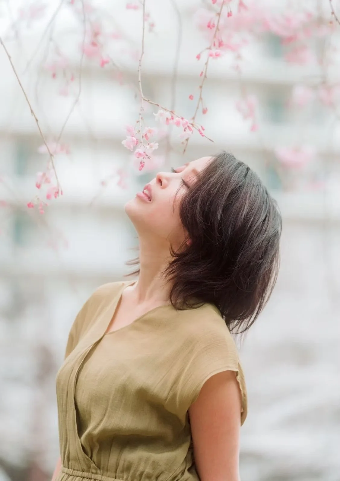 A woman with short hair stands beneath cherry blossom trees, looking up with a calm expression. The soft pink blossoms surround her, creating a peaceful, springtime atmosphere.