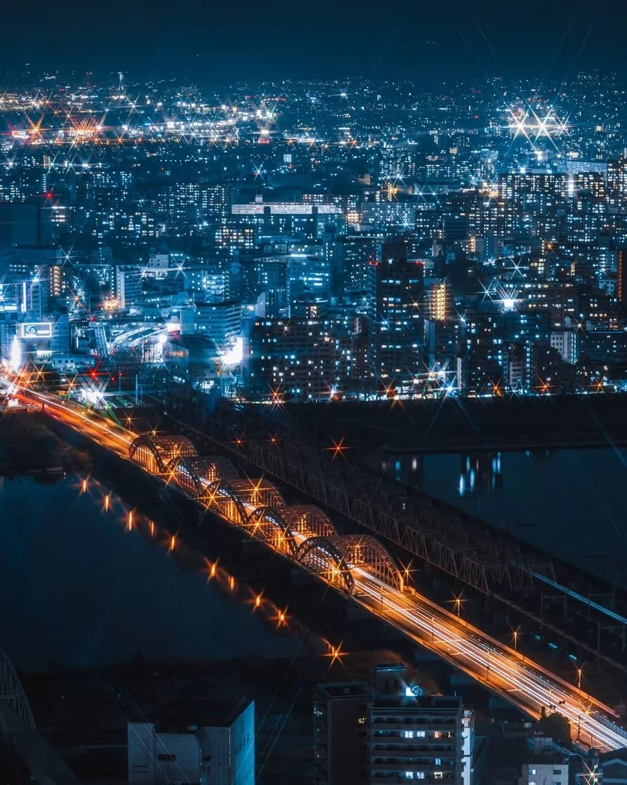Nighttime city skyline with illuminated buildings, a bridge with orange lights, and light trails from moving vehicles.