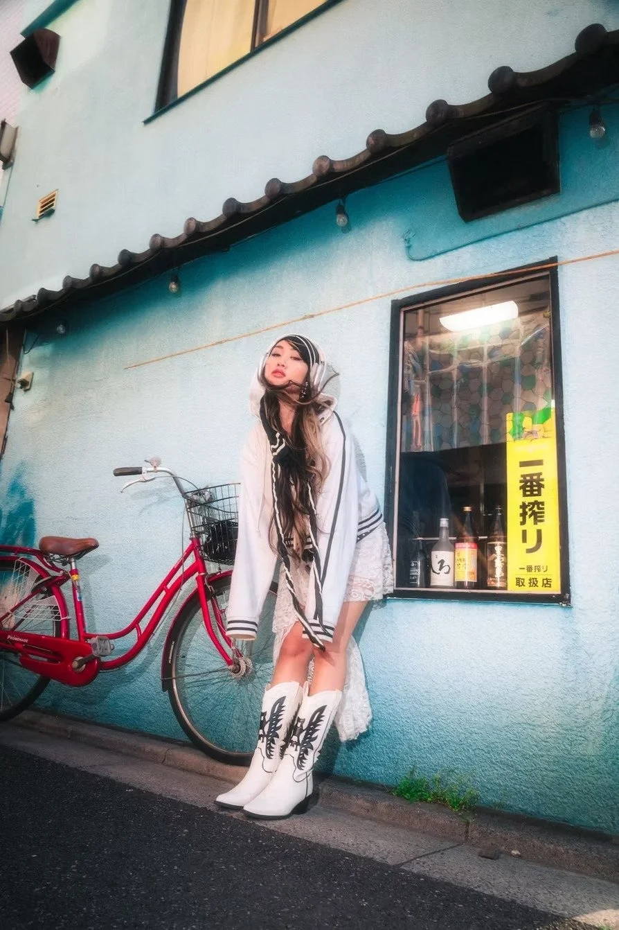 A young woman standing next to a bicycle on a city street at night, wearing white cowboy boots with black designs, lace shorts, and a white jacket with black stripes. She has long hair and a headscarf, and is leaning against a light blue wall with a 