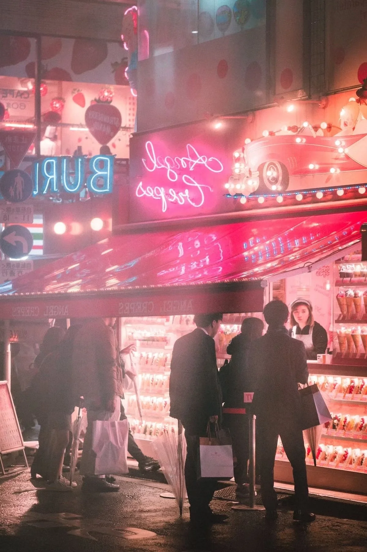 Harajuku night scene of a pink neon-lit street food stand selling ice cream and snacks, with several people ordering and walking by, illuminated by streetlights and neon signs.