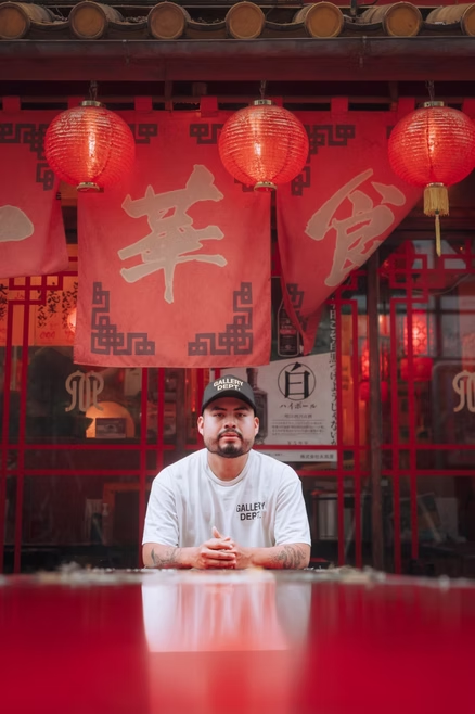 A man sitting at a red table in front of a Chinese restaurant with red lanterns and red banners with Chinese characters, wearing a cap and a white T-shirt.