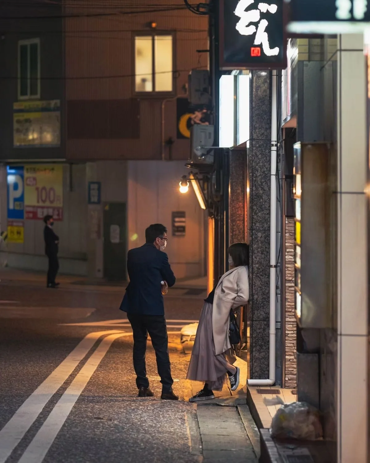 Two people stand and converse on a dimly lit street corner at night, with another person standing in the background near a building. All three are wearing face masks, with the man in a dark suit and the woman in a beige coat and long skirt.