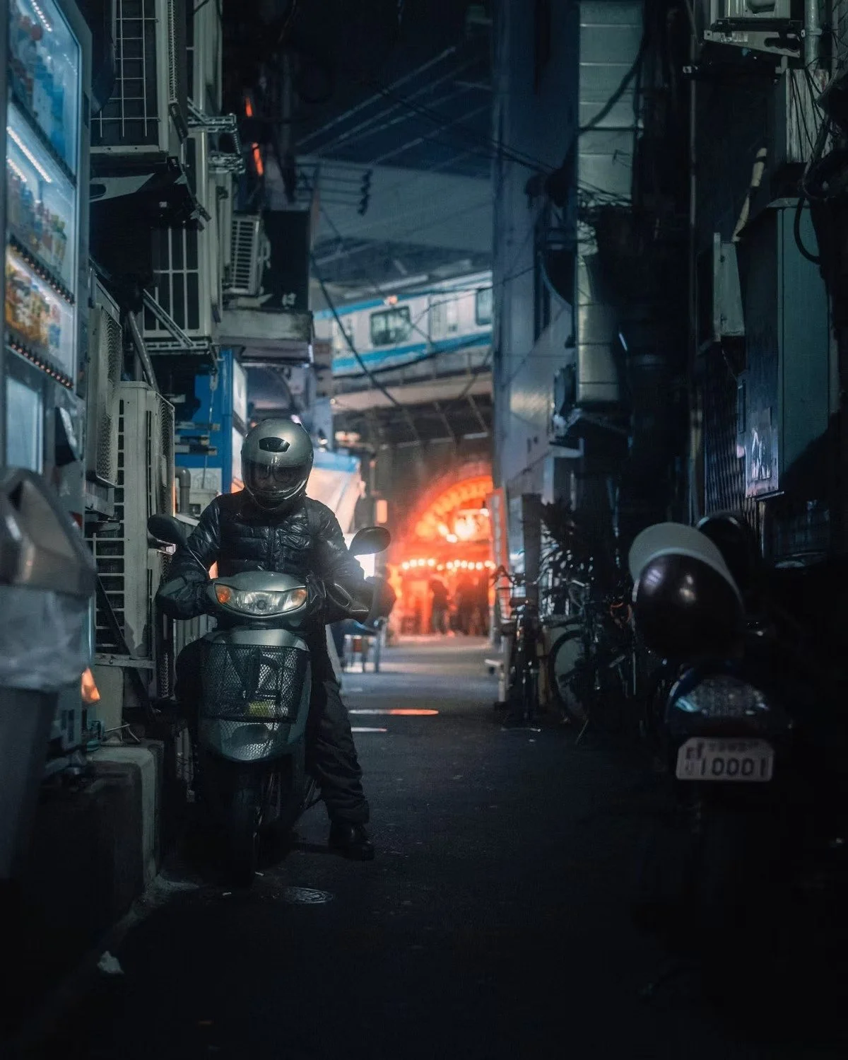 A person wearing a black helmet and jacket on a scooter in a narrow Shimbashi Tokyo alley at night, with neon signs and parked motorcycles visible.