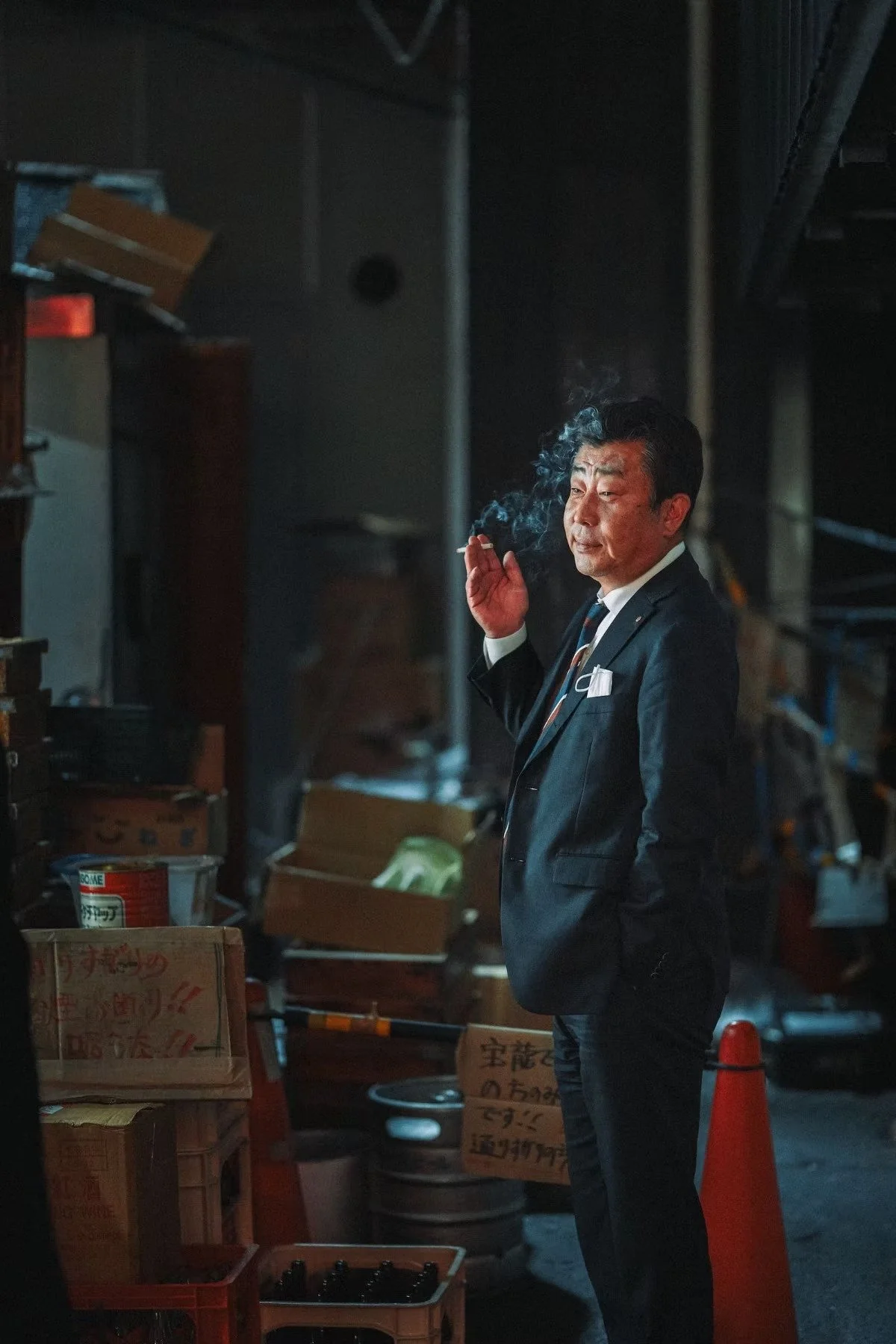 A salaryman in Tokyo a dark suit and tie stands in a dimly lit room, smoking a cigarette, surrounded by cardboard boxes and various items, with shelves and storage in the background. Captured by street photographer Deniz Demir.