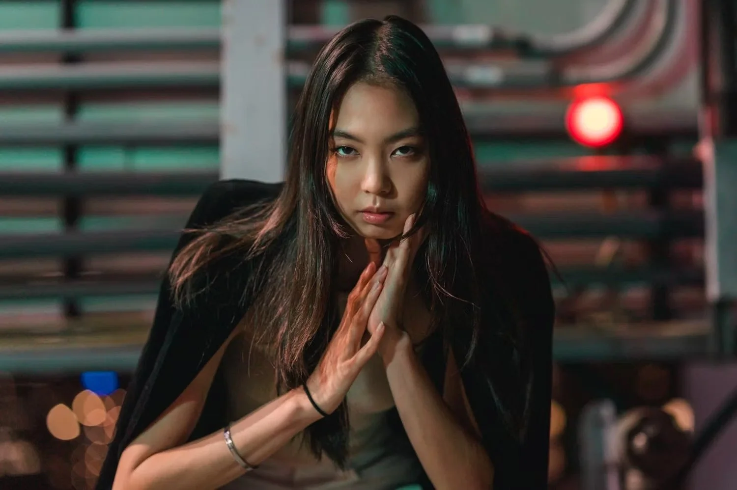 A young woman with long dark hair and intense expression posing in front of a background with horizontal lines and a red light.