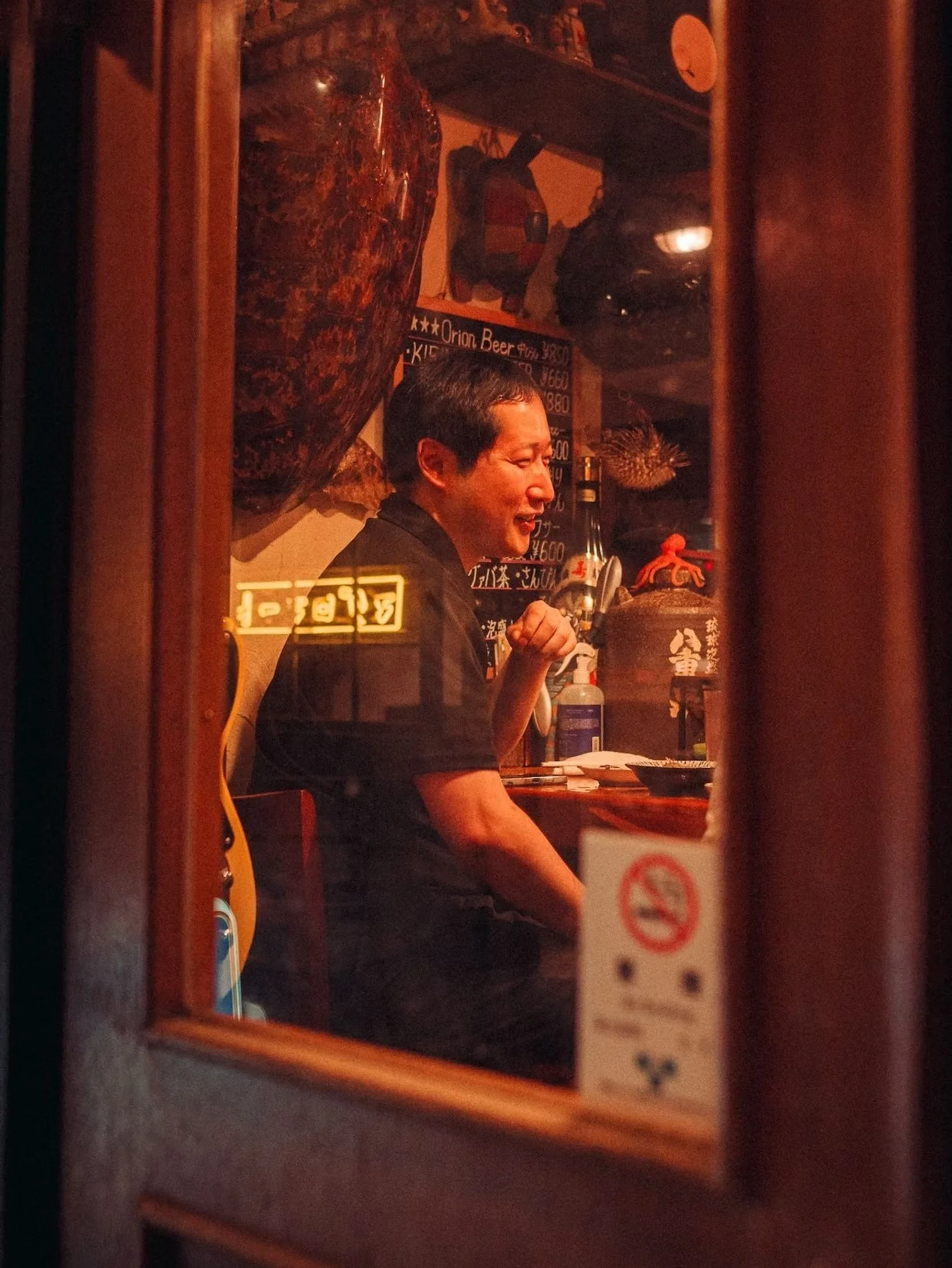 A man with dark hair sits at a counter inside a Japanese restaurant, smiling. The photo is taken through a window, with reflections and a 'No Smoking' sign partially visible in the foreground. There are various decorations and signs on the wall behind him, including a menu written in Japanese.