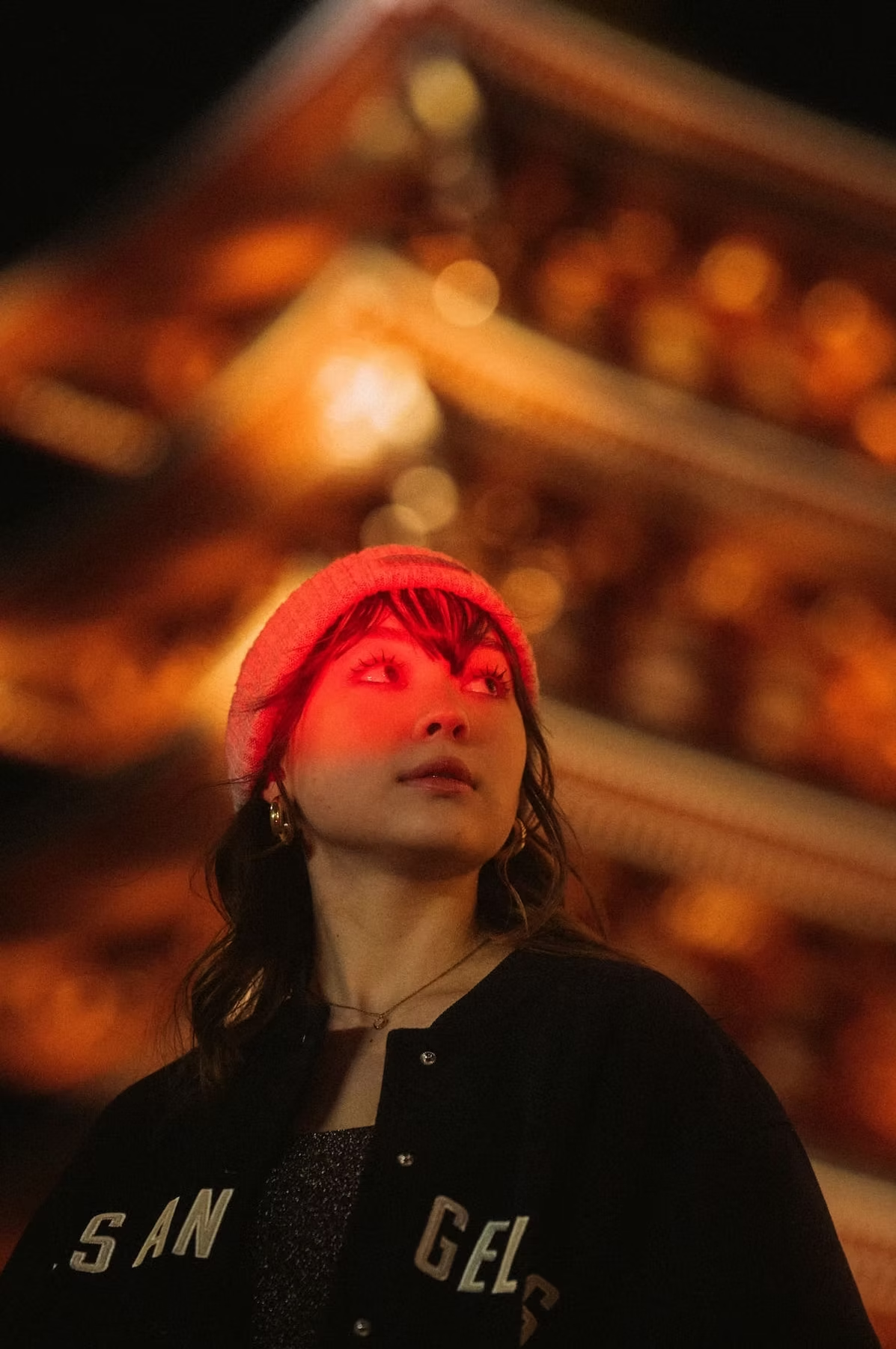 A woman wearing a black jacket and red beanie stands in front of a blurred, warm-lit background, with red lighting highlighting her face and adding dramatic effect.