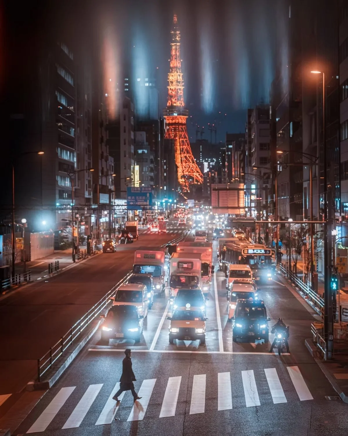 Nighttime city street with cars and a pedestrian crossing, Tokyo Tower illuminated in orange in the background, with water mist or rain creating a reflective glow.