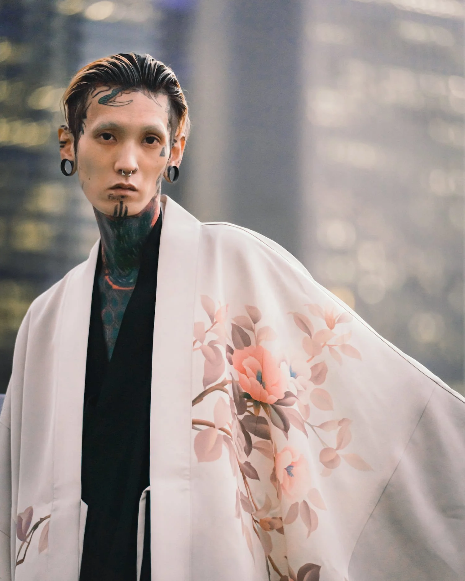 A person with tattoos and piercings wearing a light-colored kimono with a floral pattern, standing outdoors with a blurred cityscape background.