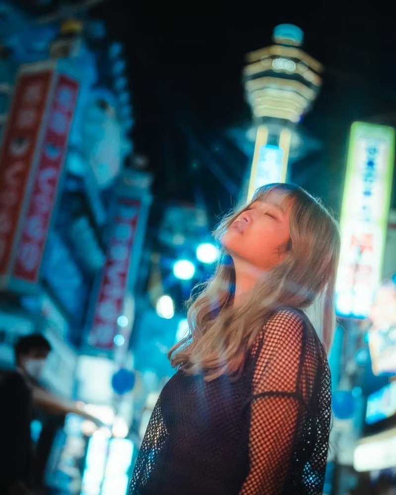 Young woman with long wavy hair, wearing a black mesh top, standing with her eyes closed, in a vibrant, brightly lit urban area at night with neon signs.