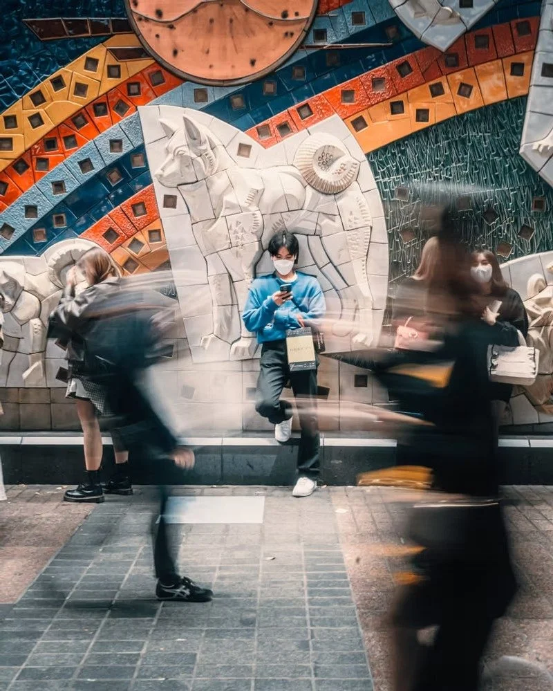 A young man wearing a mask and blue hoodie leaning against a wall with elaborate animal and geometric rock carvings, surrounded by blurred pedestrians in motion.