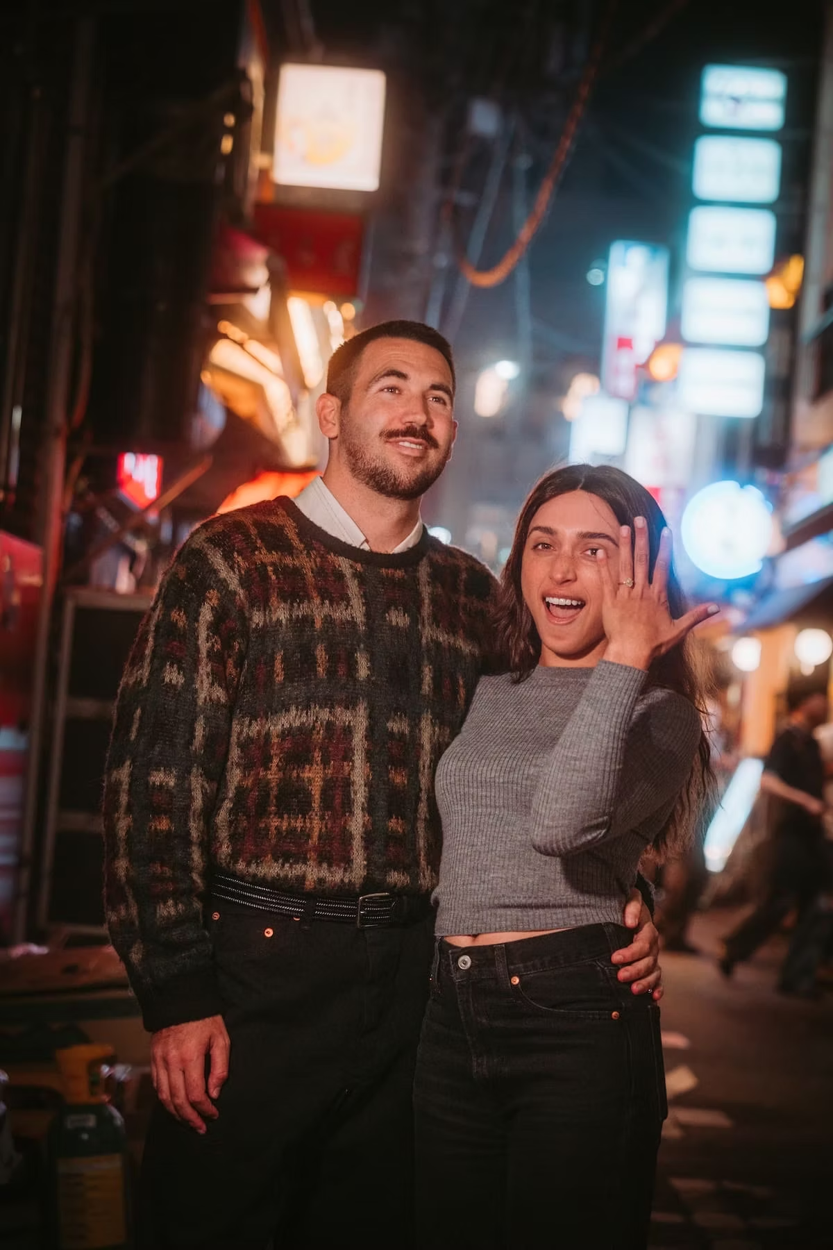 A man and woman standing together on a city street at night, smiling and posing for the camera, with bright neon signs and lights in the background.