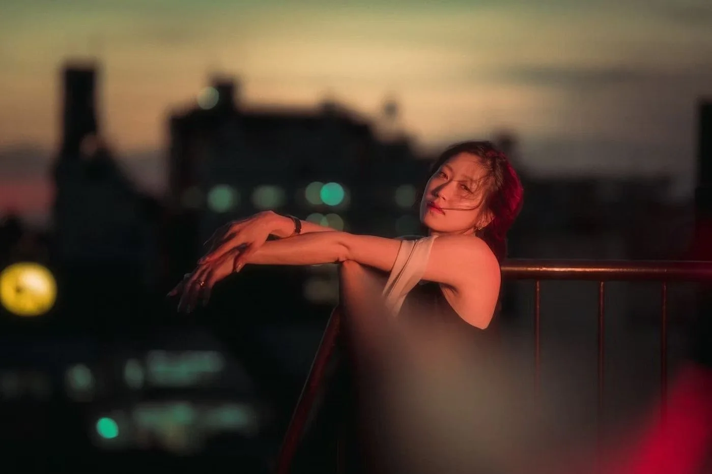 A woman with short hair leaning on a railing outdoors during dusk, with city lights in the background on a Tokyo rooftop. 