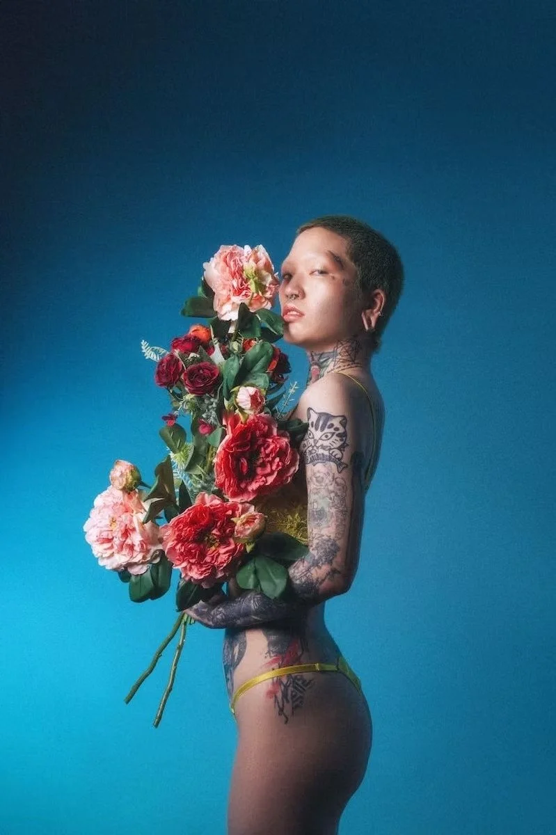 A model stands confidently while holding a bouquet of colorful flowers in a blue-lit setting. The striking colors and lighting enhance the visual impact, creating a calm, yet bold atmosphere.