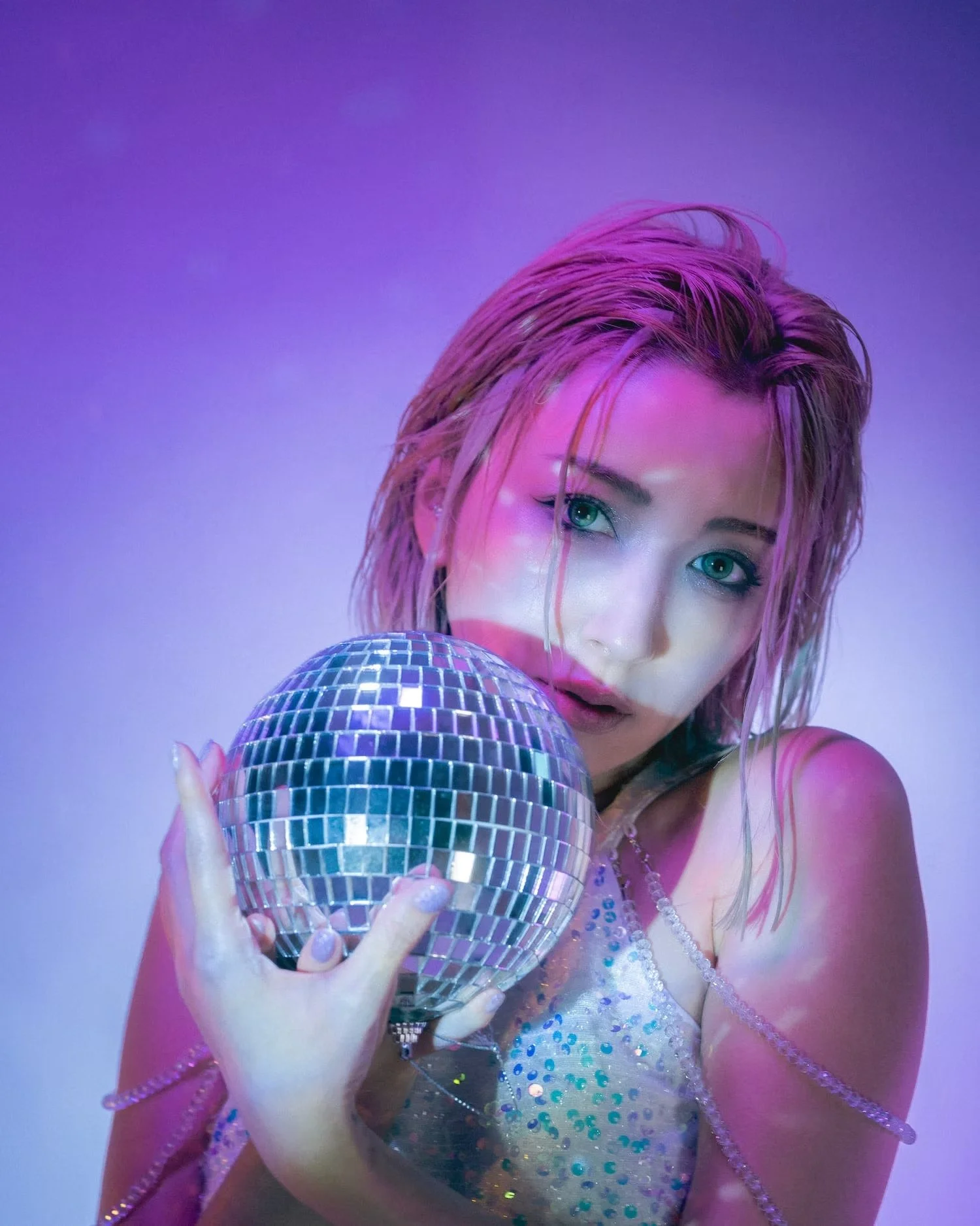 A woman with pink hair holding a small disco ball, with reflective and colorful lighting creating a vibrant atmosphere.