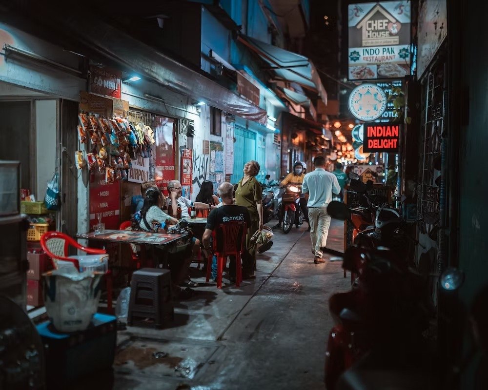 Night scene of a busy street with street food vendors, diners, and pedestrians. Signs for Indian restaurants and other eateries are visible, with motorcycles parked along the sidewalk. Shot by Tokyo street photographer Deniz Demir on the streets of S
