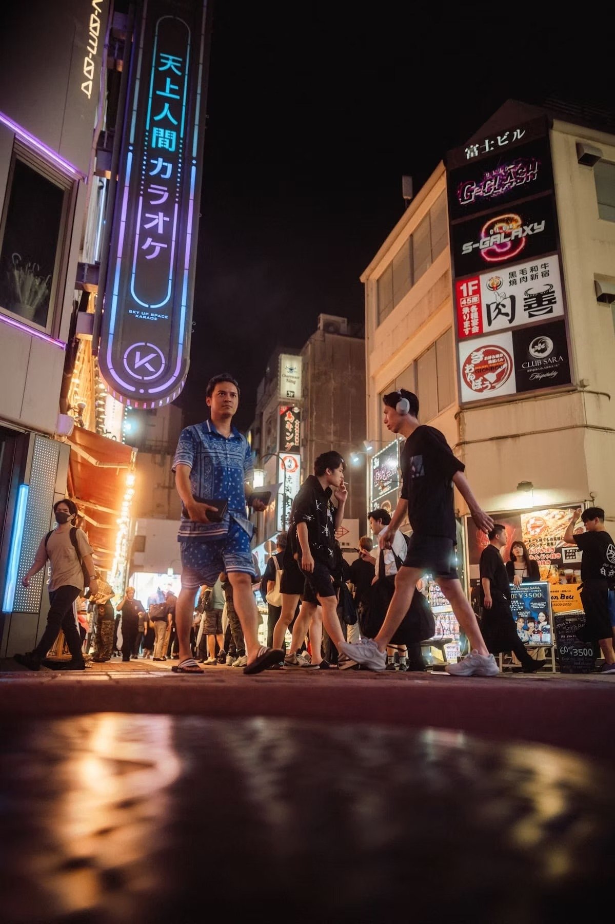 Night scene of a busy street with people walking, shopping, and socializing under neon lights and signboards, including Japanese characters, in an urban area. Kabukicho Shinjuku at night.