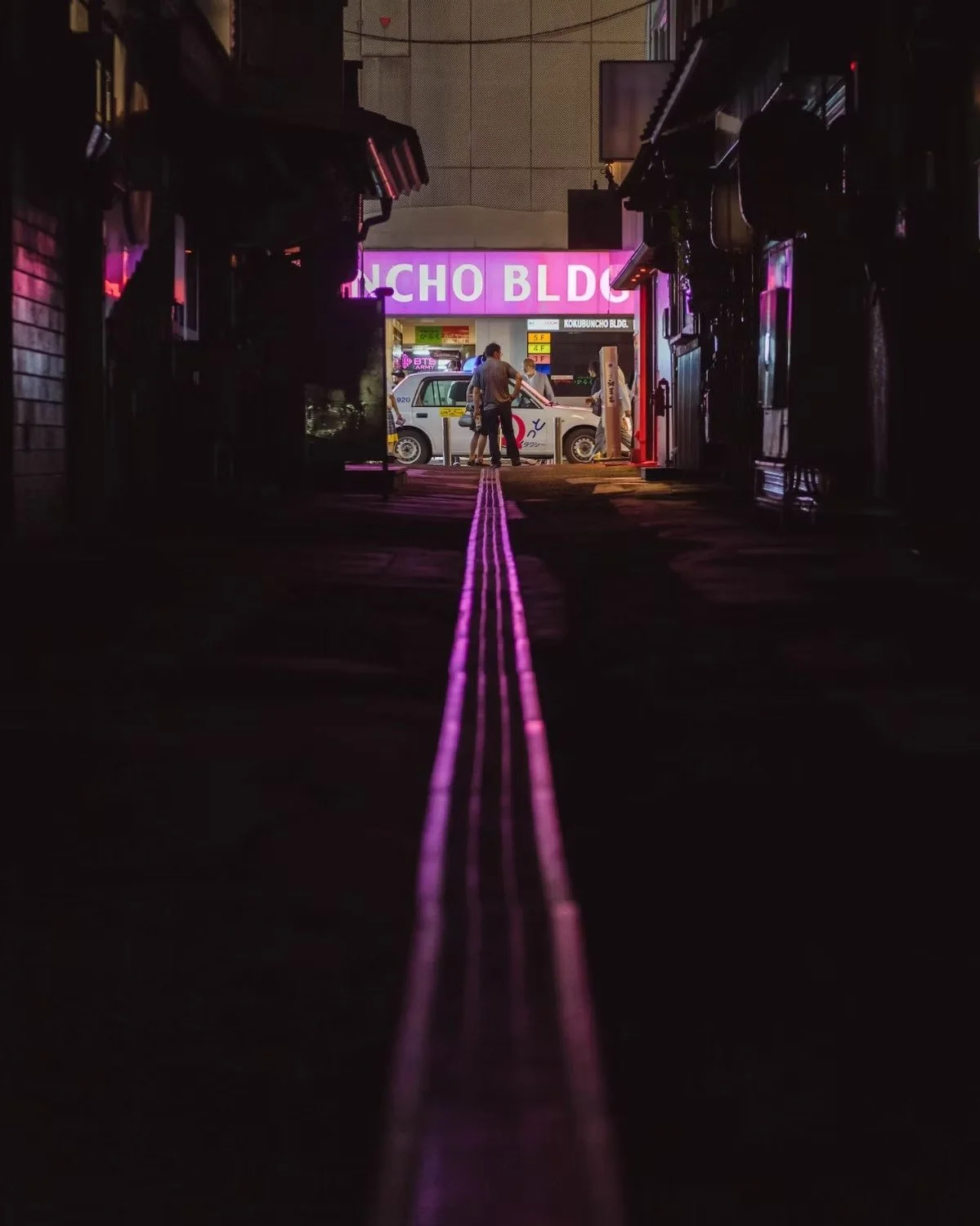 Nighttime alleyway with purple neon sign reading 'KONCHO BLDG' at the end, with people and a vehicle visible in the distance in Fukuoka.