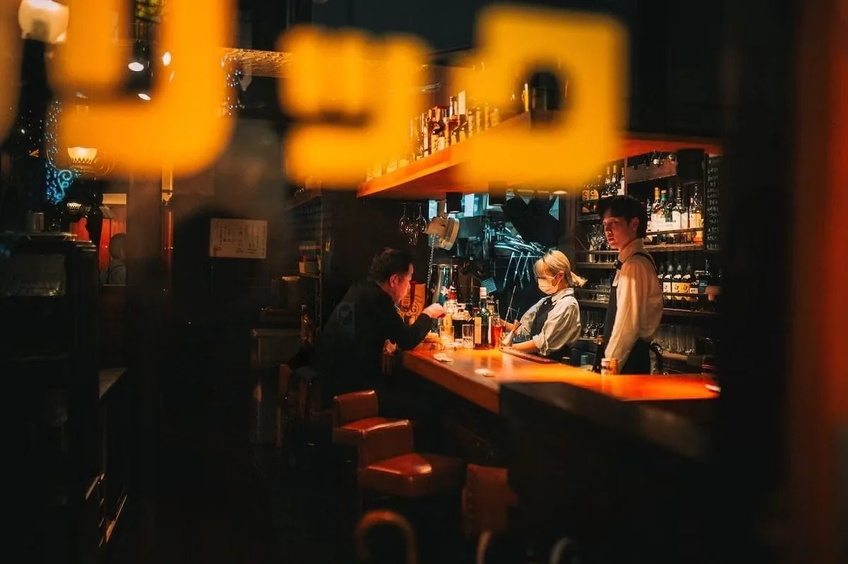 A dimly lit bar with three people sitting at the counter and a bartender attending to them, shelves filled with bottles of liquor in the background. Nakano in Tokyo night scenes.