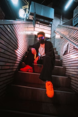 A person in a dark outfit sits on outdoor stairs in a neon-lit urban Tokyo alley, with bright lights and signs in the background.