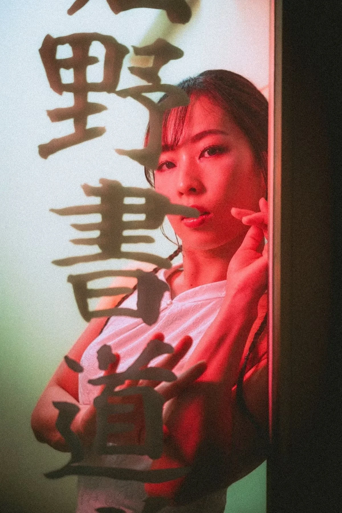Portrait photography of a Japanese woman with dark hair and makeup, looking at the camera, behind a transparent surface with black Japanese characters written on it.