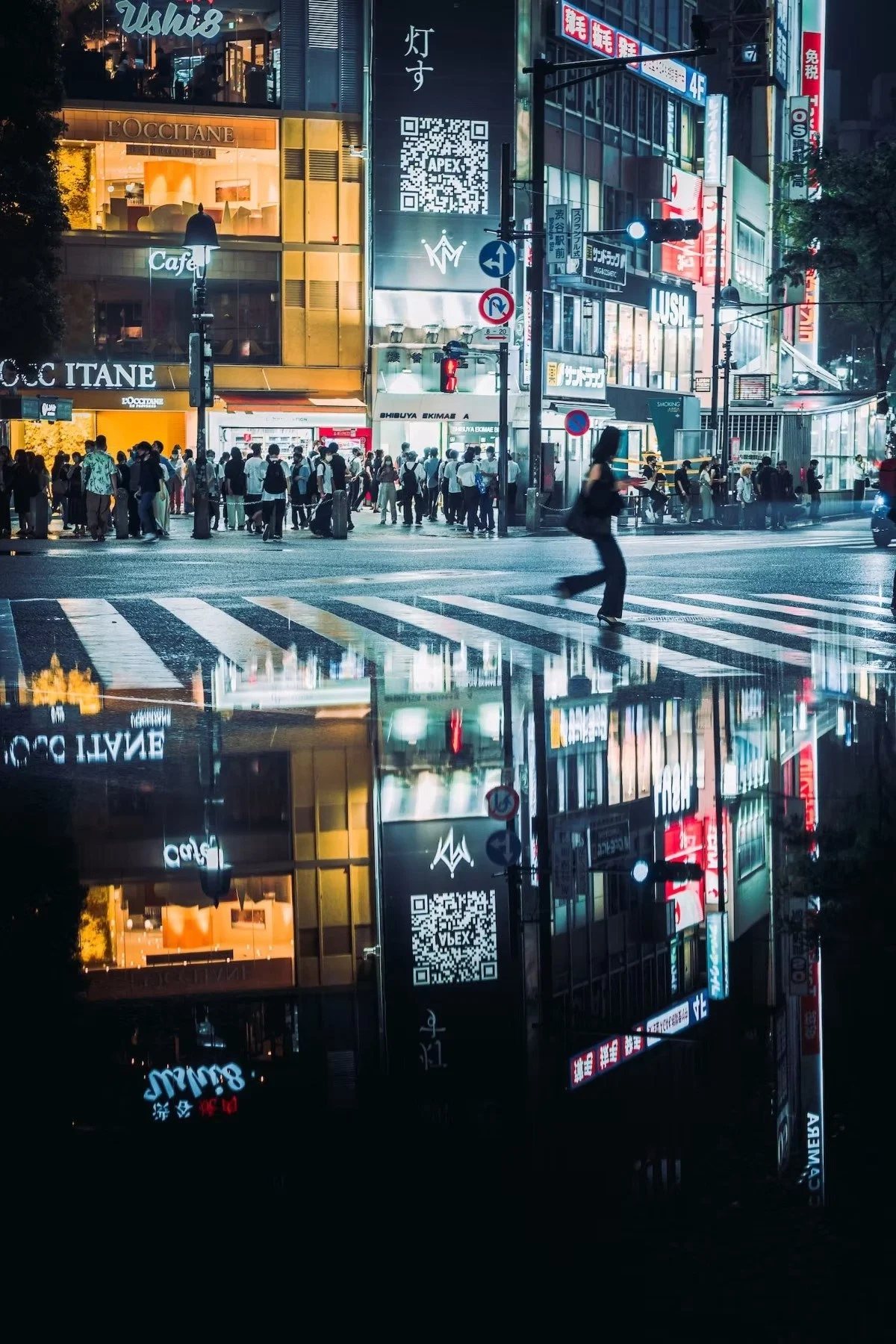 Night scene of a busy Shibuya Crossing with a crowd of pedestrians, illuminated signs, and storefronts reflecting on a wet surface, creating a mirror effect.