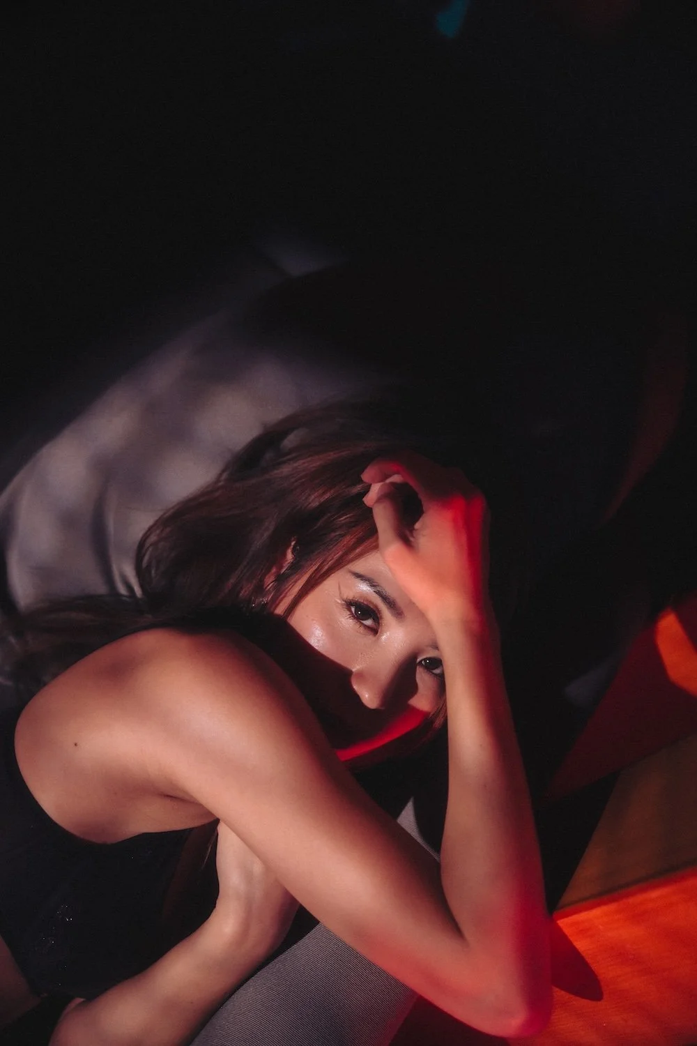 An asian woman with long brown hair, lying on her side in a dark environment with red cinematic lighting, resting her head on her arm and gazing at the camera in a seductive boudoir pose in a hotel.