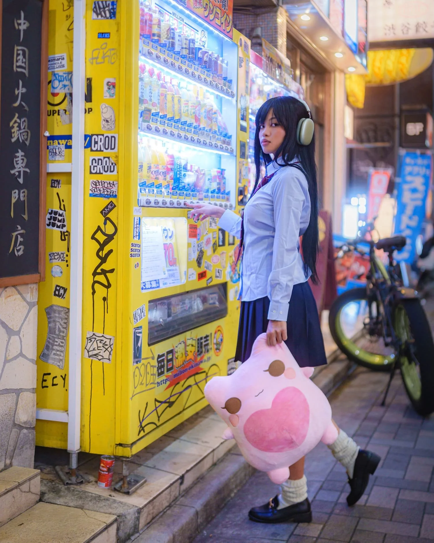 A young woman with long black hair wearing a school uniform, headphones, and black shoes is leaning against a yellow vending machine on a city sidewalk. She is holding a pink plush toy with a cute face. The background features bicycles and illuminated signs.