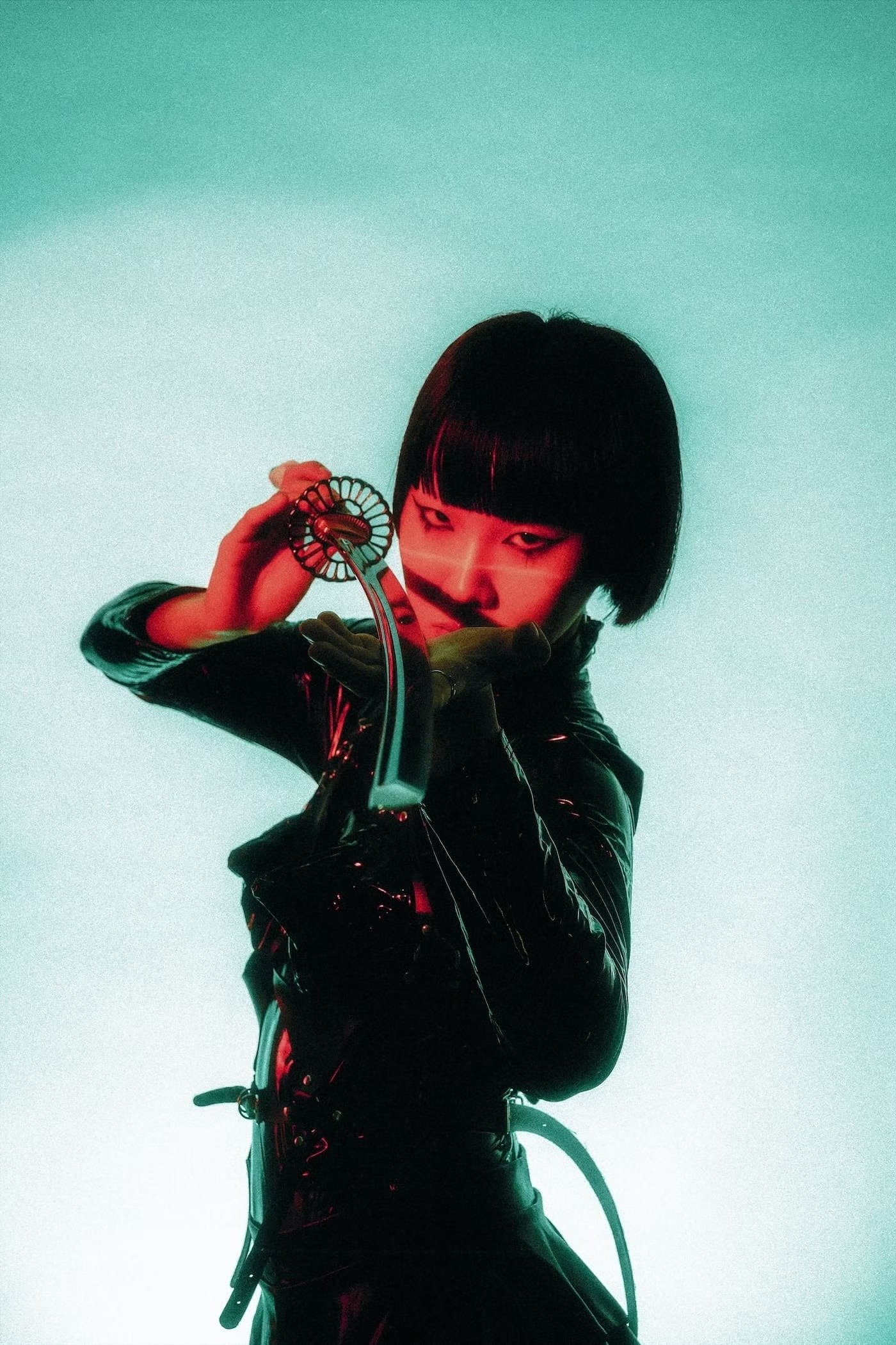 A woman with short black hair and a serious expression holding a curved knife with a decorative handle, wearing a shiny black jacket, against a gradient background of teal to white. Studio portrait photography by Deniz Demir.