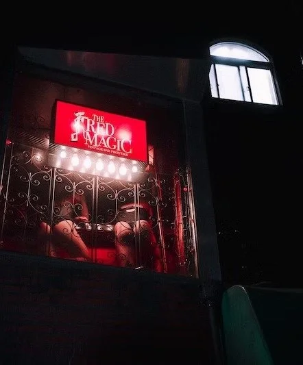 A stage with a red sign that reads 'The Forest of Magic' and is lit by bright stage lights. The stage is enclosed by decorative wrought iron bars, and there is a window with an arched top in a dark room.