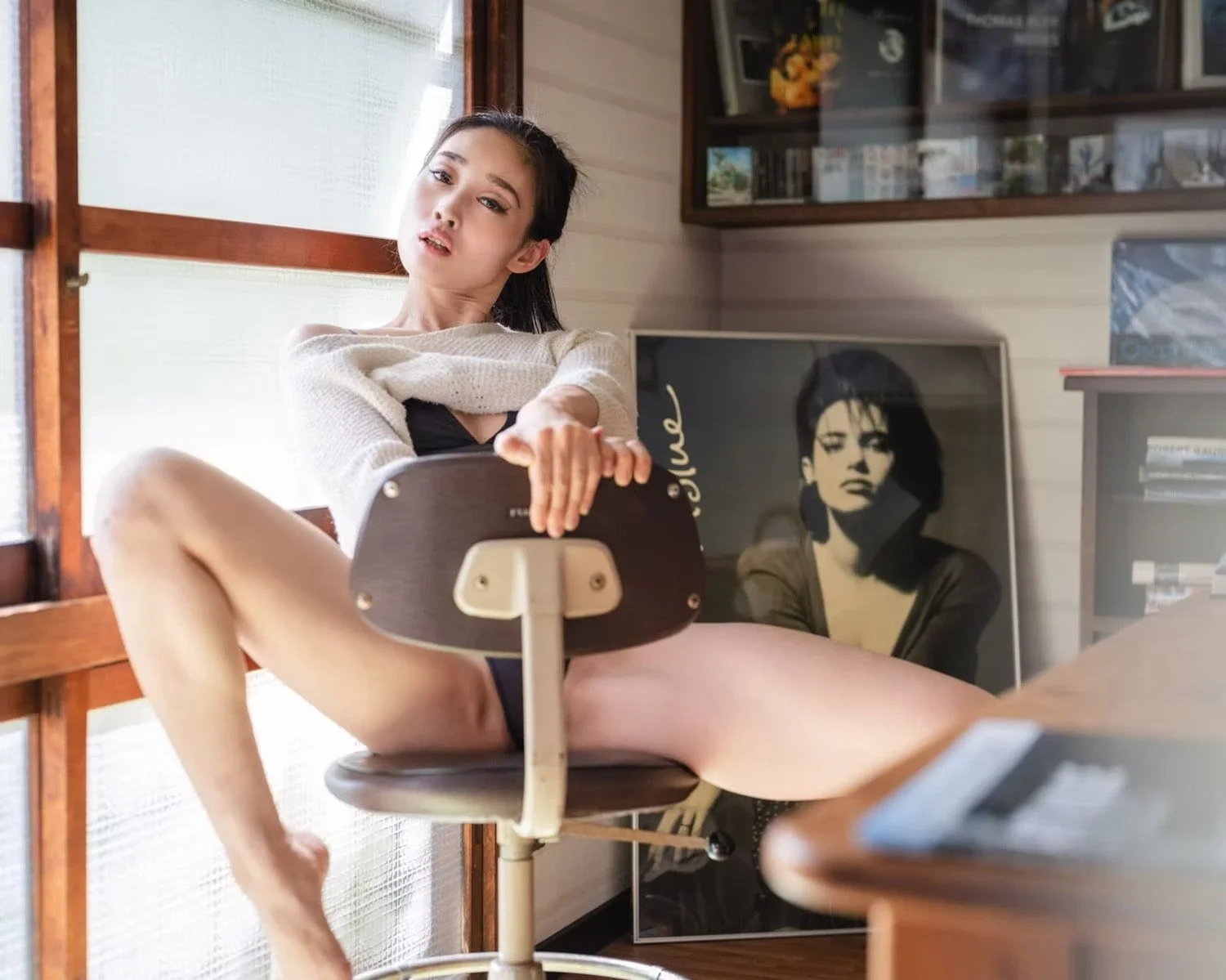 A woman sitting on a chair with her legs apart, wearing a beige sweater and black underwear, in a room with wooden walls and large windows, with a large portrait of a woman on the floor behind her.