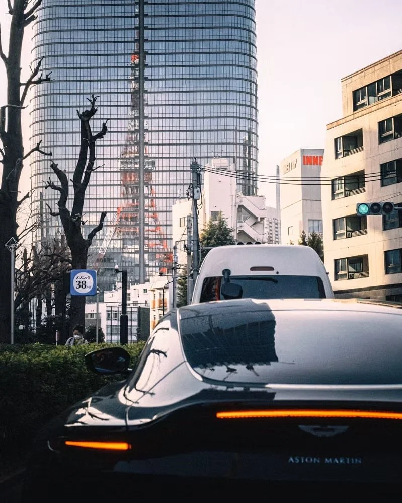 City street scene with high-rise buildings, a reflective glass skyscraper showing Tokyo Tower, a leafless tree, a white van, and a black Aston Martin sports car in the foreground. Some pedestrians are visible.