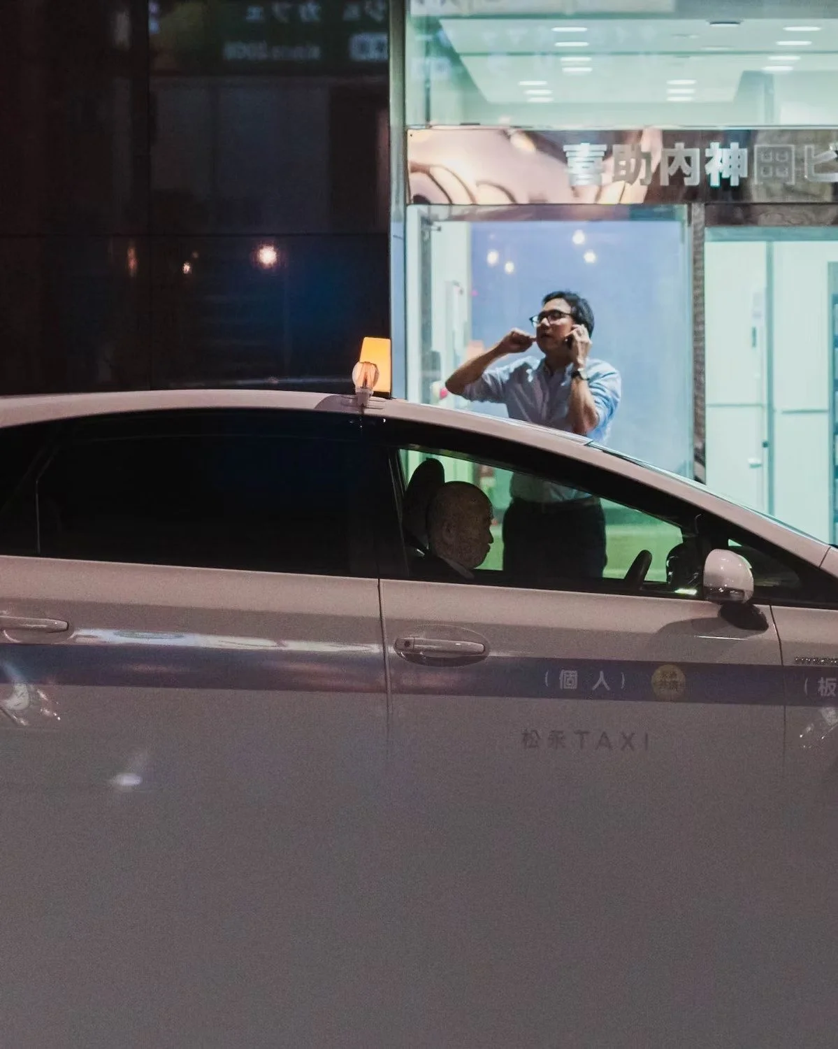 A taxi parked outside a building at night. The driver, an elderly man, is sitting inside the taxi, and a man in glasses, wearing a light-colored shirt, stands in a building behind a glass window talking on a mobile phone. The building has Japanese ch