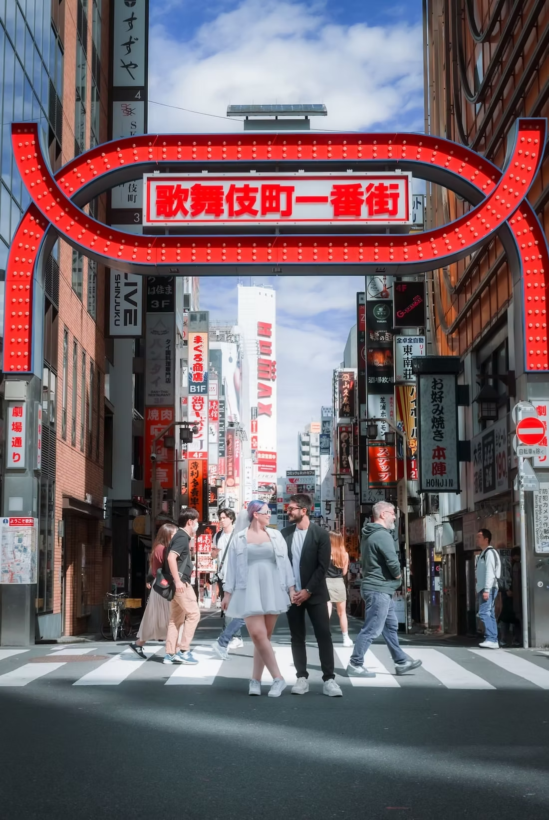 A busy street in the Kabukicho area of Tokyo, with neon signs and advertisements in the background. People walk across the crosswalk beneath a large red archway, representing the iconic entrance to the entertainment district.