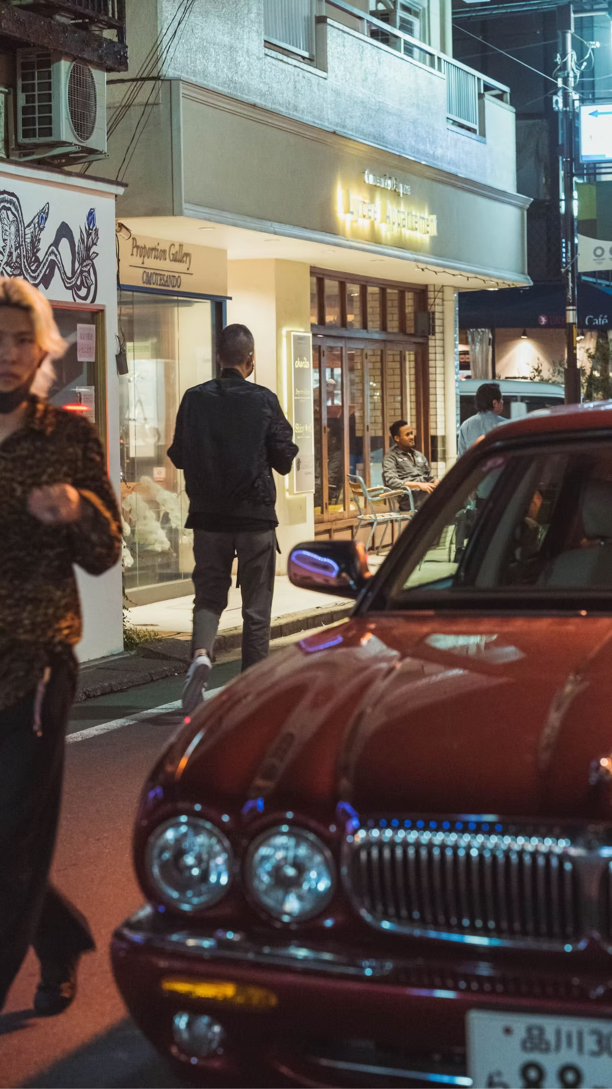 Nighttime street scene with a maroon vintage car in the foreground, multiple people walking and sitting outside a restaurant or café, illuminated storefronts, and a sign that says 'Licea Aguacate'.