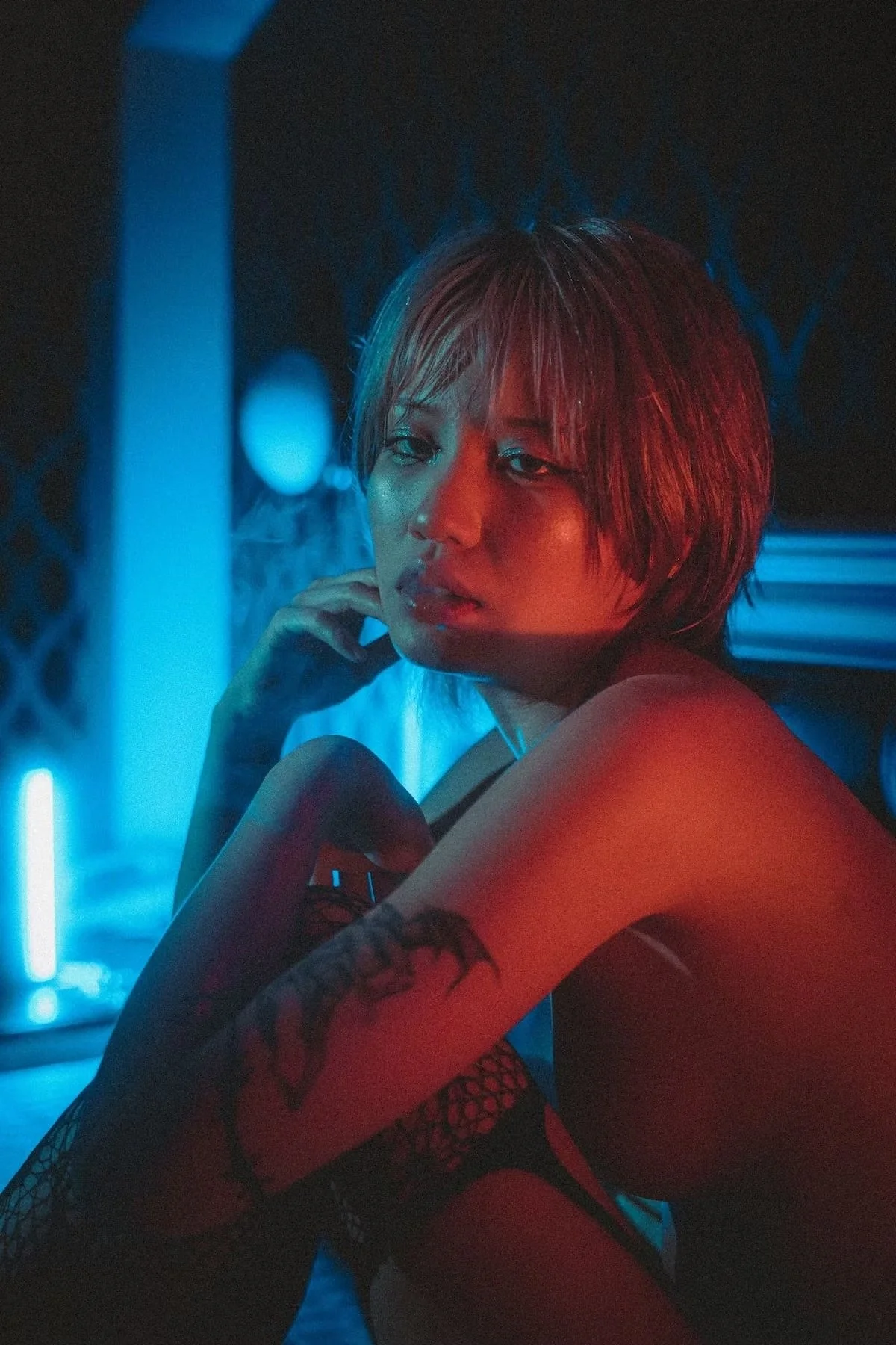 A woman with short hair and tattoos on her arm, sitting in a dimly lit space with blue and red lighting, resting her chin on her hand and looking at the camera.