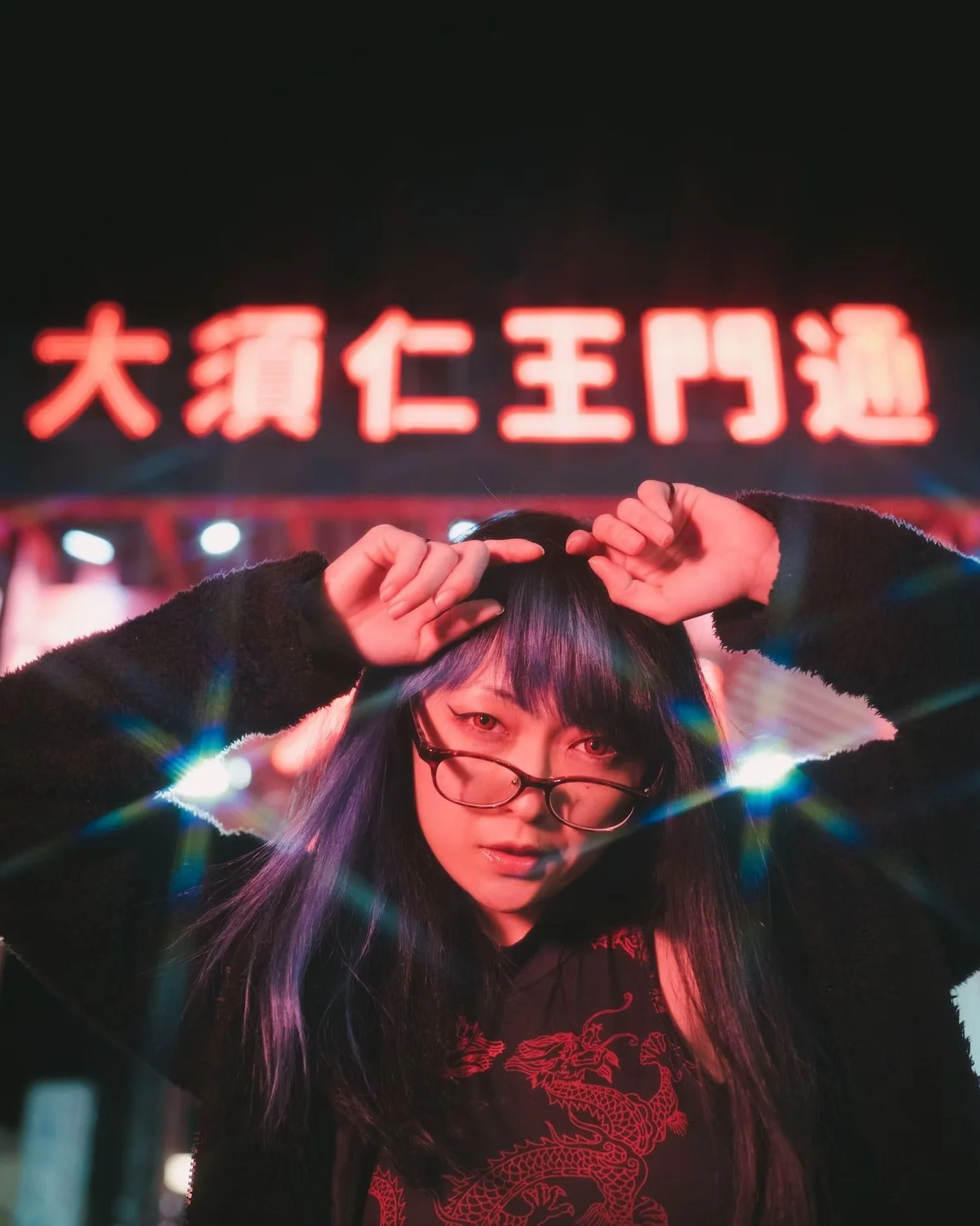A young woman with glasses and dark hair, with colored highlights, looking at the camera and posing with her hands near her head, standing in front of a brightly lit red neon sign with Chinese characters.